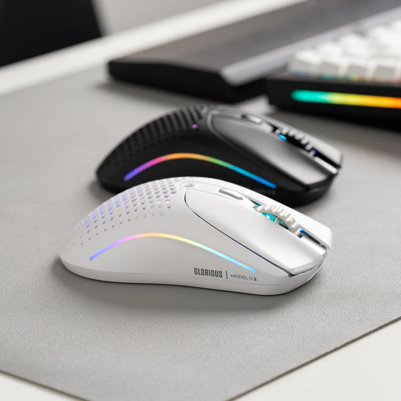 A white Glorious Model O 2 Wireless gaming mouse with RGB lighting is placed in front of a black Model O 2 Wireless on a mousepad.